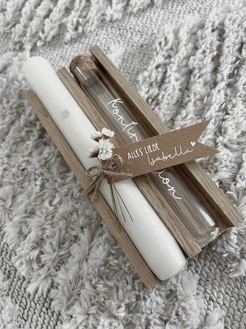 Puede incluir: Una vela blanca y un vial de vidrio con la palabra "Konfir" se muestran en un soporte de madera. Se adjunta una peque&ntilde;a etiqueta con las palabras "Alles Liebe Isabella". Decorado con flores secas.