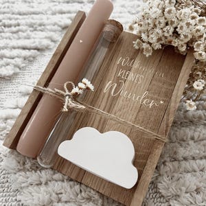 Puede incluir: Una pieza decorativa de madera con una vela rosa, un vial de vidrio y un adorno blanco en forma de nube. La pieza de madera tiene grabadas las palabras "Willkommen kleines Wunder". Los artículos están atados con hilo y decorados con pequeñas flores blancas.