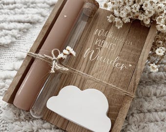 May include: A wooden decorative piece with a pink candle, a glass vial, and a white cloud-shaped ornament. The wooden piece has the words "Willkommen kleines Wunder" engraved on it. The items are tied together with twine and decorated with small white flowers.