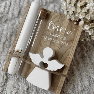 Puede incluir: Una placa de madera con el nombre "Emma" y detalles de nacimiento. Incluye una vela blanca, un tubo de vidrio y una figura de ángel blanco. La placa está atada con hilo y adornada con pequeñas flores blancas.