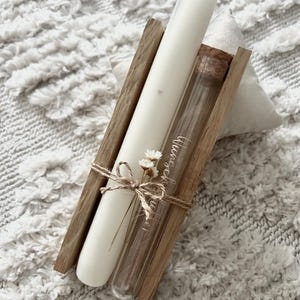 Puede incluir: Un juego decorativo de velas y viales de vidrio. La vela marfil tiene un pequeño detalle de corazón y se sostiene en un marco de madera con un vial de vidrio. El vial está sellado con un corcho y atado con hilo y pequeñas flores.