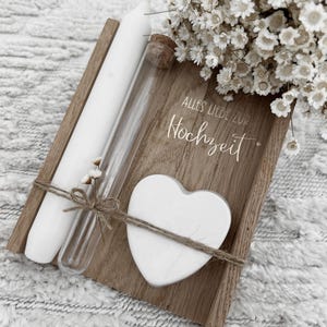 Puede incluir: Una bandeja de madera con una vela blanca, un vial de vidrio y un corazón blanco atado con hilo. La bandeja tiene el texto alemán "Alles Liebe zur Hochzeit". Un ramo de flores blancas en el fondo. Sobre una superficie blanca texturizada.