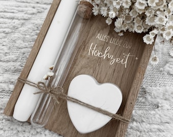 May include: A wooden tray with a white candle, a glass vial, and a white heart tied with twine. The tray has the German text "Alles Liebe zur Hochzeit". A bouquet of white flowers is in the background. The items are arranged on a white textured surface.