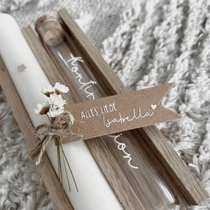 Puede incluir: Una vela blanca y un vial de vidrio con tap&oacute;n de corcho se presentan en una caja de madera. El vial tiene texto blanco. Un peque&ntilde;o ramo de flores blancas y una etiqueta de papel marr&oacute;n con texto blanco est&aacute;n atados a la vela.