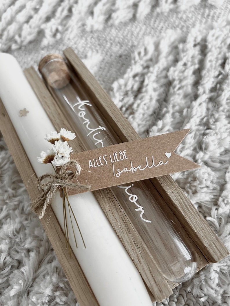 Puede incluir: Una vela blanca y un vial de vidrio con tap&oacute;n de corcho se presentan en una caja de madera. El vial tiene texto blanco. Un peque&ntilde;o ramo de flores blancas y una etiqueta de papel marr&oacute;n con texto blanco est&aacute;n atados a la vela.