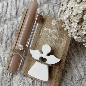 May include: A wooden plaque with the text "ALLES LIEBE ZUR KOMMUNION" is adorned with a white angel figurine, a brown candle, and a glass vial. The items are tied together with twine and small white flowers.