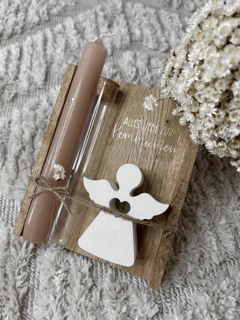 May include: A wooden plaque with the text "ALLES LIEBE ZUR KOMMUNION" is adorned with a white angel figurine, a brown candle, and a glass vial. The items are tied together with twine and small white flowers.