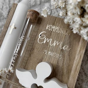May include: A white communion candle with a silver cross, a glass vial with a cork stopper, and a wooden box with the name "Emma" and the date "25. Mai 2025" engraved on it. A white angel figurine is attached to the box.