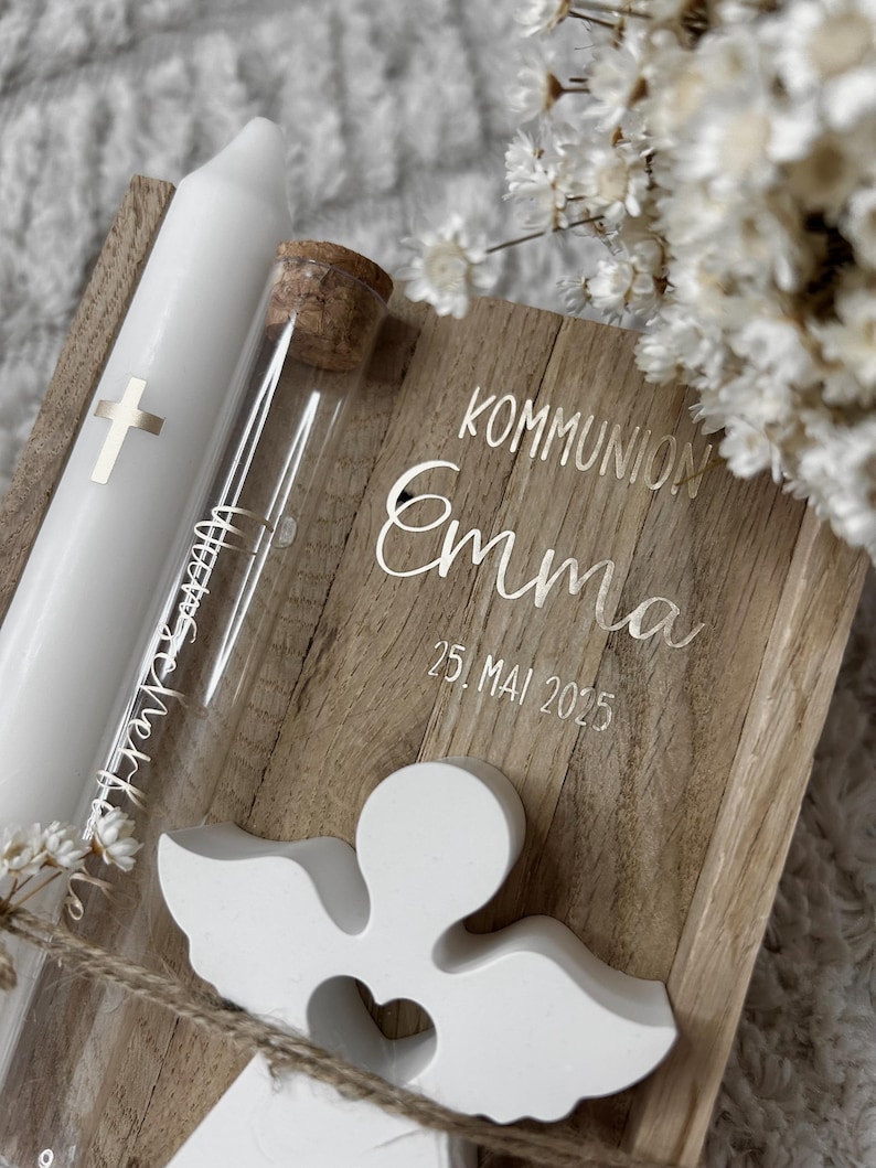 May include: A white communion candle with a silver cross, a glass vial with a cork stopper, and a wooden box with the name "Emma" and the date "25. Mai 2025" engraved on it. A white angel figurine is attached to the box.