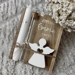 May include: A wooden keepsake box with the text "KOMMUNION Emma 25. MAI 2025" and a white angel cutout. Includes a white candle with a cross and a glass vial with a cork stopper, tied together with twine. A bouquet of white flowers is in the background.