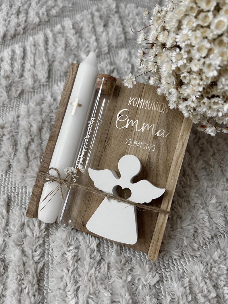May include: A wooden keepsake box with the text "KOMMUNION Emma 25. MAI 2025" and a white angel cutout. Includes a white candle with a cross and a glass vial with a cork stopper, tied together with twine. A bouquet of white flowers is in the background.