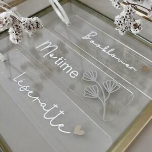 Puede incluir: Marcapáginas transparentes de acrílico con texto blanco y diseños florales. Los marcapáginas dicen "Me time", "Booklover" y "Leseratte". Cada marcapáginas tiene una cinta blanca y un pequeño corazón. Se muestran en una caja de cristal con marco dorado.