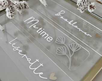 May include: Clear acrylic bookmarks with white text and floral designs. The bookmarks read "Me time", "Booklover", and "Leseratte". Each bookmark has a white ribbon and a small heart accent. The bookmarks are displayed in a gold-framed glass box.