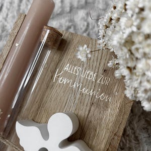 May include: A wooden plaque with the text "ALLES LIEBE ZUR KOMMUNION" is accompanied by a white angel figurine, a glass tube with a cork, and a pink candle. White flowers are in the background.