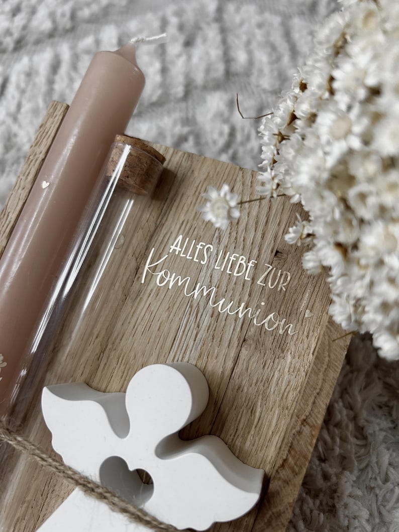 May include: A wooden plaque with the text "ALLES LIEBE ZUR KOMMUNION" is accompanied by a white angel figurine, a glass tube with a cork, and a pink candle. White flowers are in the background.