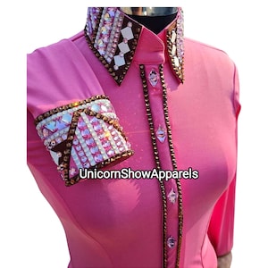 May include: Pink long-sleeve show shirt with brown and white rhinestone embellishments on the collar and cuffs. The shirt has a button-up front and the text "UnicornShowApparels" is printed on the front.