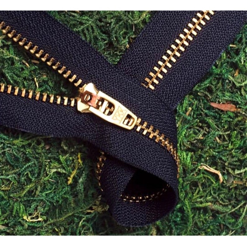 May include: A close-up of a black zipper with a gold metal slider and teeth. The zipper is lying on a bed of green moss.