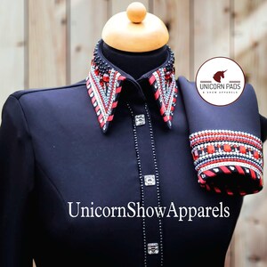 May include: Black show shirt with a red and white embellished collar and cuffs. The collar has a red ribbon trim and the cuffs have a black and white striped pattern. The shirt is decorated with rhinestones and the text "UnicornShowApparels" is printed on the front. The text "Unicorn Pads & Show Apparels" is printed on a white circle with a unicorn logo.