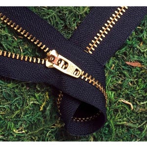 May include: A close-up of a black zipper with a gold metal slider and teeth. The zipper is lying on a bed of green moss.