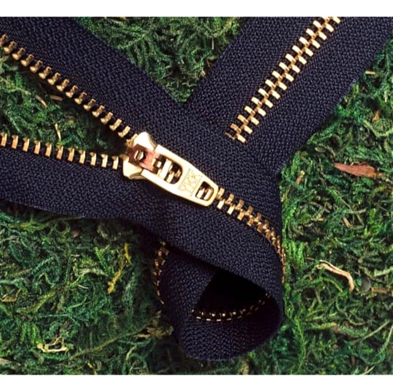 May include: A close-up of a black zipper with a gold metal slider and teeth. The zipper is lying on a bed of green moss.