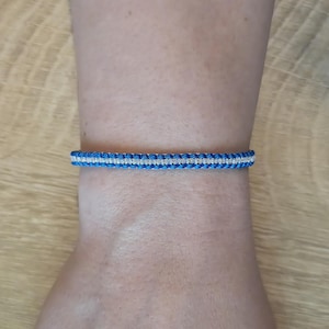 Op de afbeelding: Een blauw en zilver geweven armband. De armband is een band van blauwe draad met een dunne zilveren streep in het midden. De armband wordt om een pols gedragen.