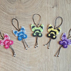 Może przedstawiać: Pięć makramowych ozdób w kształcie motyli w kolorach różowym, niebieskim, zielonym, żółtym i fioletowym. Każdy motyl ma czarną pętlę do zawieszenia i zwisające czarne sznurki z białymi koralikami. Motyle ułożone są na jasnobrązowej drewnianej powierzchni.