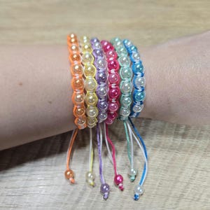 Op de afbeelding: Een verzameling kleurrijke kralenarmbanden. Elke armband heeft een rij imitatieparels in tinten oranje, geel, paars, roze en blauw. De armbanden zijn vastgebonden met bijpassende gekleurde koorden, wat een levendig accessoire creëert.