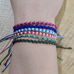 Op de afbeelding: Een verzameling kleurrijke vriendschapsarmbanden in verschillende tinten roze, blauw, wit, rood, groen en olijfgroen. De armbanden zijn gemaakt van geknoopt draad en worden op een pols gestapeld. De achtergrond is een lichtbruin houten oppervlak.