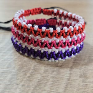 Op de afbeelding: Een stapel handgemaakte armbanden met kralen in rood, wit, roze en paars. Elke armband heeft een uniek patroon van kralen en geweven draad. De armbanden zijn op elkaar gestapeld en tonen de verschillende kleurencombinaties en ontwerpen.