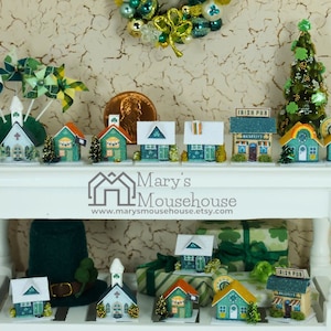 May include: A white table with two shelves displays a collection of miniature houses decorated in green, white, and gold. The houses feature shamrocks, a pub sign, and a church steeple. The table is decorated with a green wreath, a green Christmas tree, and a green hat.