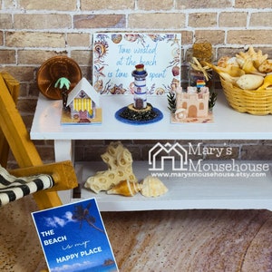 May include: Miniature beach scene with a white table, featuring a striped beach chair, a sign that reads "The beach is my happy place", and various beach-themed miniatures, including a lighthouse and a sandcastle. A basket of seashells is also present.