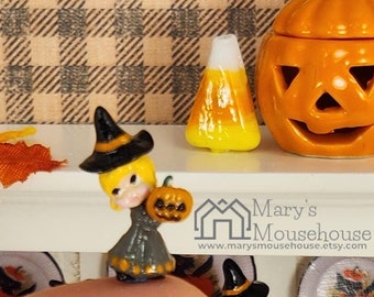 May include: A miniature white hutch with shelves displaying Halloween-themed decorations. There are several miniature witches holding pumpkins, a small ceramic pumpkin, and a sign that reads "Cute as a Pumpkin".