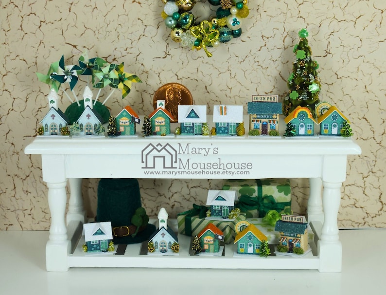 May include: A white table with two shelves displays a collection of miniature houses decorated in green, white, and gold. The houses feature shamrocks, a pub sign, and a church steeple. The table is decorated with a green wreath, a green Christmas tree, and a green hat.