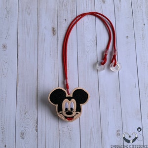 May include: A Mickey Mouse face mask lanyard with a red cord and clear plastic adjusters. The embroidered Mickey Mouse face is black, tan, white, and red. The lanyard is on a light wood grain background.