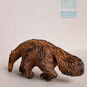 Puede incluir: Figurita detallada de un oso hormiguero de madera, que muestra un hocico largo y un cuerpo marrón texturizado con marcas marrones más oscuras. La figura está posada en una postura de caminar, capturando las características distintivas del animal.