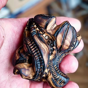 Puede incluir: Escultura de madera tallada a mano con un acabado marrón oscuro. La obra de arte presenta formas y texturas orgánicas intrincadas, posiblemente representando una criatura o forma vegetal abstracta. La pieza se sostiene en una mano, mostrando su tamaño y artesanía.