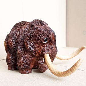 May include: A detailed wooden sculpture of a woolly mammoth. The figurine is primarily brown with textured fur and light-colored tusks. The mammoth is depicted in a standing position, showcasing its large size and prehistoric features.