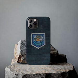 May include: A dark blue phone case with a textured appearance. The case features a colorful emblem with a sun, birds, and text. The phone case is resting on a gray stone surface.