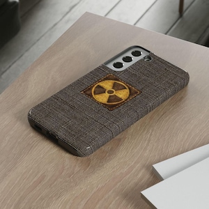 May include: A brown phone case with a textured fabric design. The case features a square patch with a yellow and brown radiation symbol. The case is designed for a smartphone.