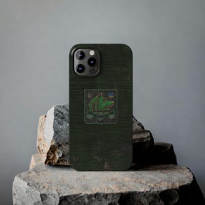 Può includere: Custodia per telefono verde scuro con finitura strutturata. La custodia presenta una toppa con un disegno di lupo verde, una bandiera gialla e blu e la scritta "BOAR". La custodia del telefono è appoggiata su una pietra grigia.
