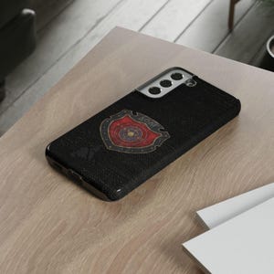 May include: Black phone case with a shield design. The shield is red and gold with the Greek letters ΔΟΛΓ. The case is on a light brown surface.