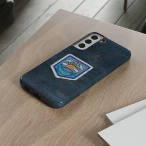 May include: A navy blue phone case with a textured appearance. The case features a white and blue emblem with a sun, waves, and text. The phone case is resting on a light brown wooden surface.