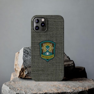 May include: A dark green phone case with a textured surface. The case features a patch with a yellow atom design and the word "MOHOЛИТ" in white. The phone case is resting on a rock.