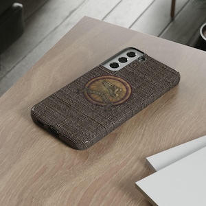May include: A brown phone case with a textured fabric design. The case features a circular emblem with a detailed illustration of a winged creature in gold and brown tones. The case is on a light-coloured wooden surface.