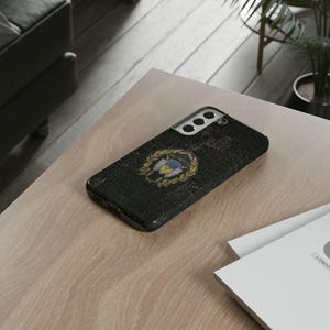 May include: Black phone case with a textured surface and a gold emblem featuring a shield and laurel wreath. The case is resting on a light-colored wooden surface, with a hint of a plant in the background.