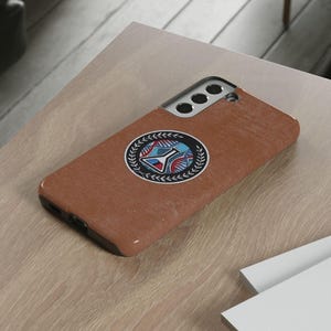 Peut inclure: Coque de téléphone marron avec un emblème décoratif représentant un motif bleu et rouge dans une couronne de laurier. L'étui est posé sur une surface en bois de couleur claire.