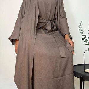 以下が含まれることがあります： 薄い茶色の3点セットの服装。セットには、長袖のトップス、スカート、それに合うロングカーディガンが含まれています。トップスはウエストにリボンが付いています。スカートはAラインで足首まであります。カーディガンは前開きで、長袖です。
