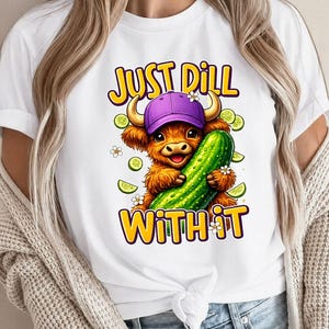 May include: White t-shirt featuring a cartoon illustration of a Highland cow wearing a purple cap and holding a large green pickle. The text "JUST DILL WITH IT" is above and below the cow.