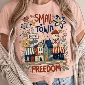 Könnte beinhalten: Hellrosa T-Shirt mit einem farbenfrohen Grafikdesign einer Kleinstadt-Szene. Das Design enthält die Worte "SMALL TOWN FREEDOM", Gebäude, Blumen und einen Wasserturm. Die Nummer "240988" ist ebenfalls auf einem Schild zu sehen.