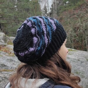 Könnte beinhalten: Eine schwarze, blaue und lila gestrickte Mütze mit einem wirbelnden Muster. Die Mütze wird von einer Person mit langen braunen Haaren getragen.